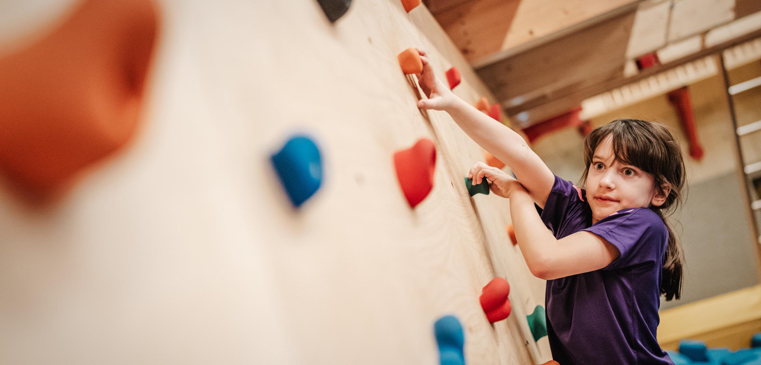 M&auml;dchen klettert an einer bunten Kletterwand mit verschiedenen Griffen in einer Turnhalle.