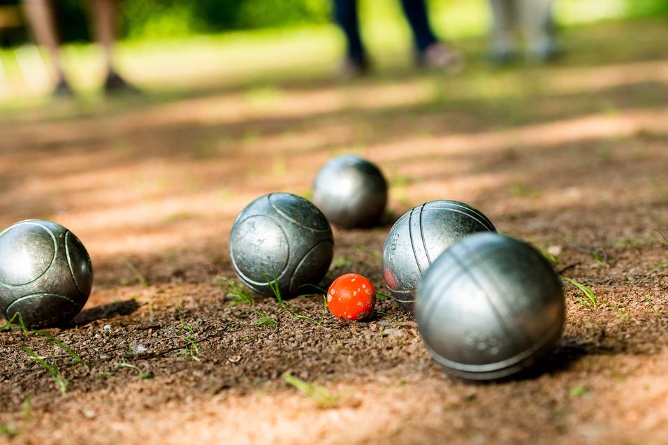 Gleise mit silbernen Boulekugeln und einer roten Kugel auf einem erdigen Boden in einem Park.