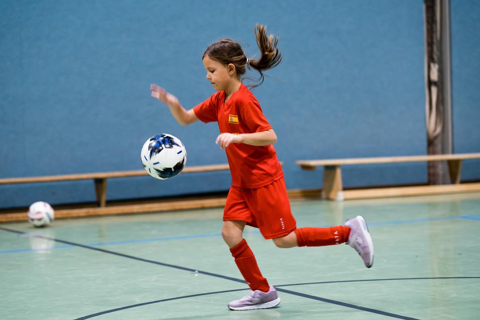 Mädchen in rotem Trikot dribbelt einen Fußball in einer Halle mit grünem Boden und blauen Wänden.