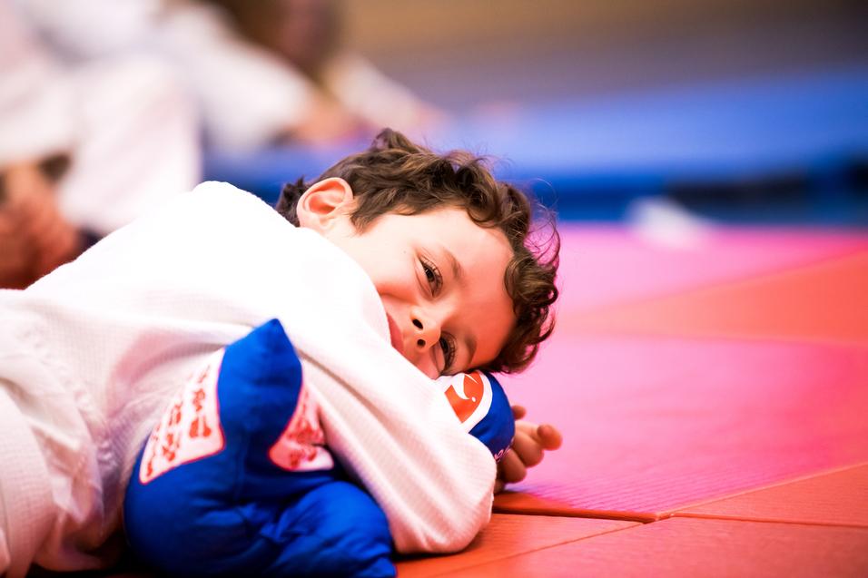Lächelnder Junge in Judoanzug, der auf einer roten Matte mit einem blauen Kissen liegt.