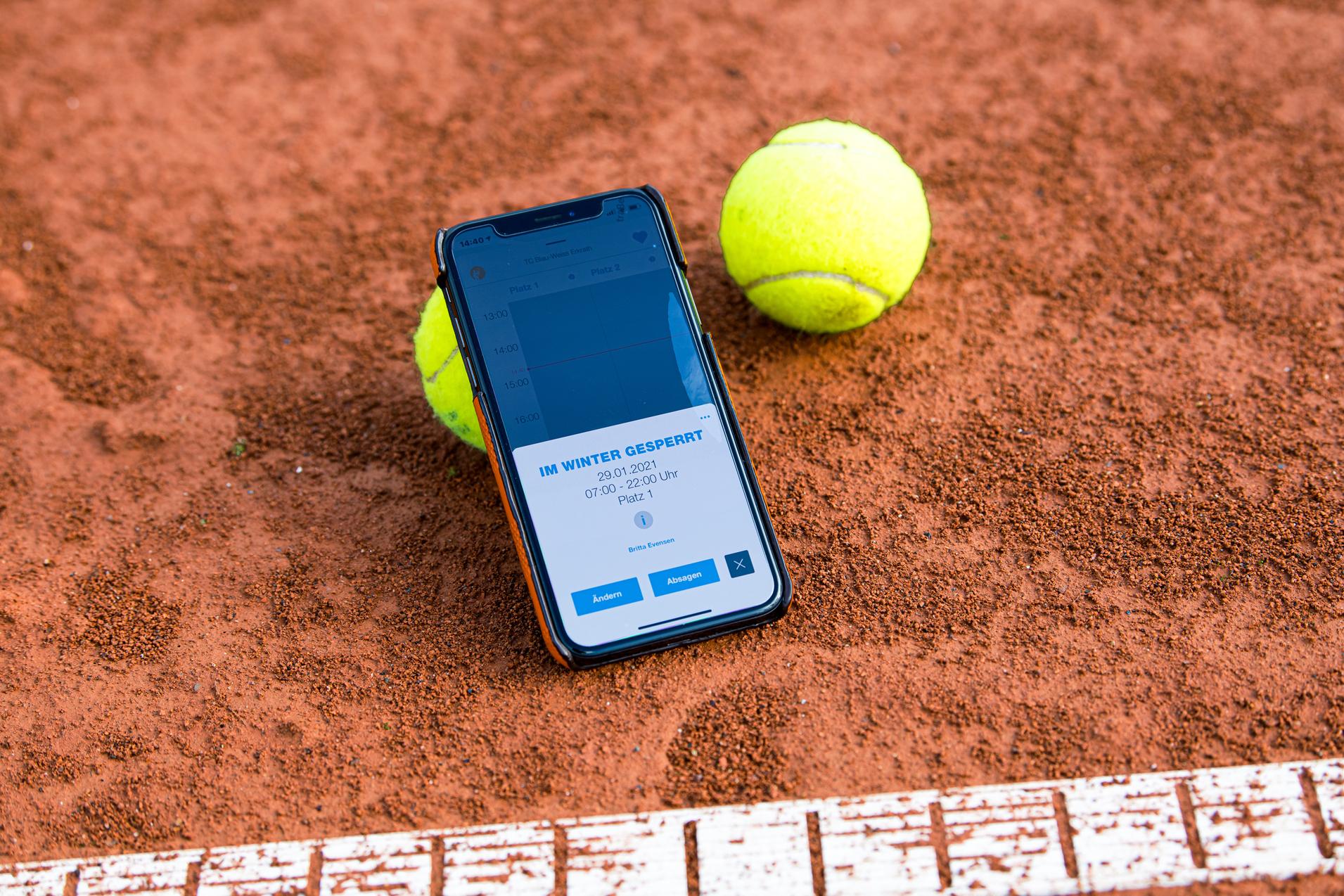 Smartphone mit Buchungsinfo liegt auf einem Tennisplatz, neben zwei gelben Tennisbällen auf rotem Sand.