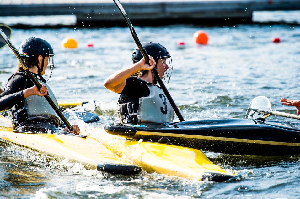 Zwei Kayakfahrerinnen paddeln auf einem Gewässer, Spritzer fliegen durch die Luft, orangefarbene Bojen sind im Hintergrund sichtbar.